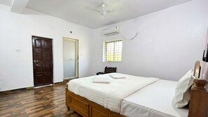 A bedroom with a wooden bed, a ceiling fan, an air conditioning unit, a window with blinds, and a door. - Celeste Stays (Candolim)