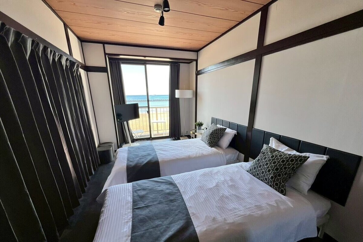 A hotel room with two beds, a large window with a view of the beach, and a ceiling fan.
