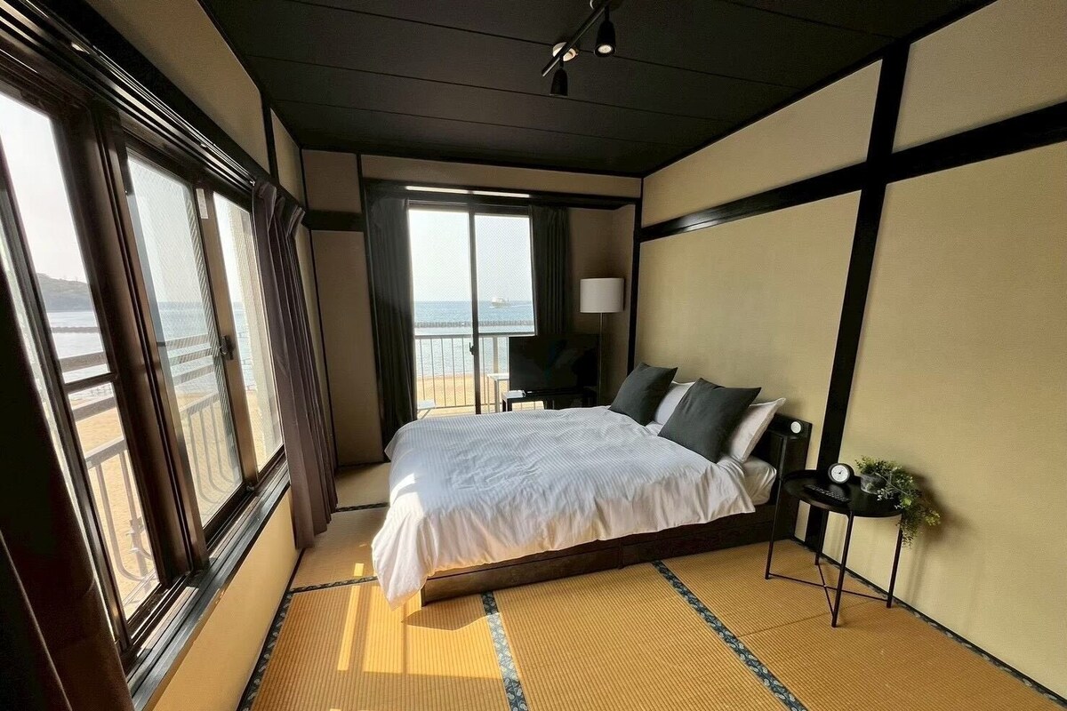 A hotel room with a bed, a nightstand, a lamp, and a view of the ocean.
