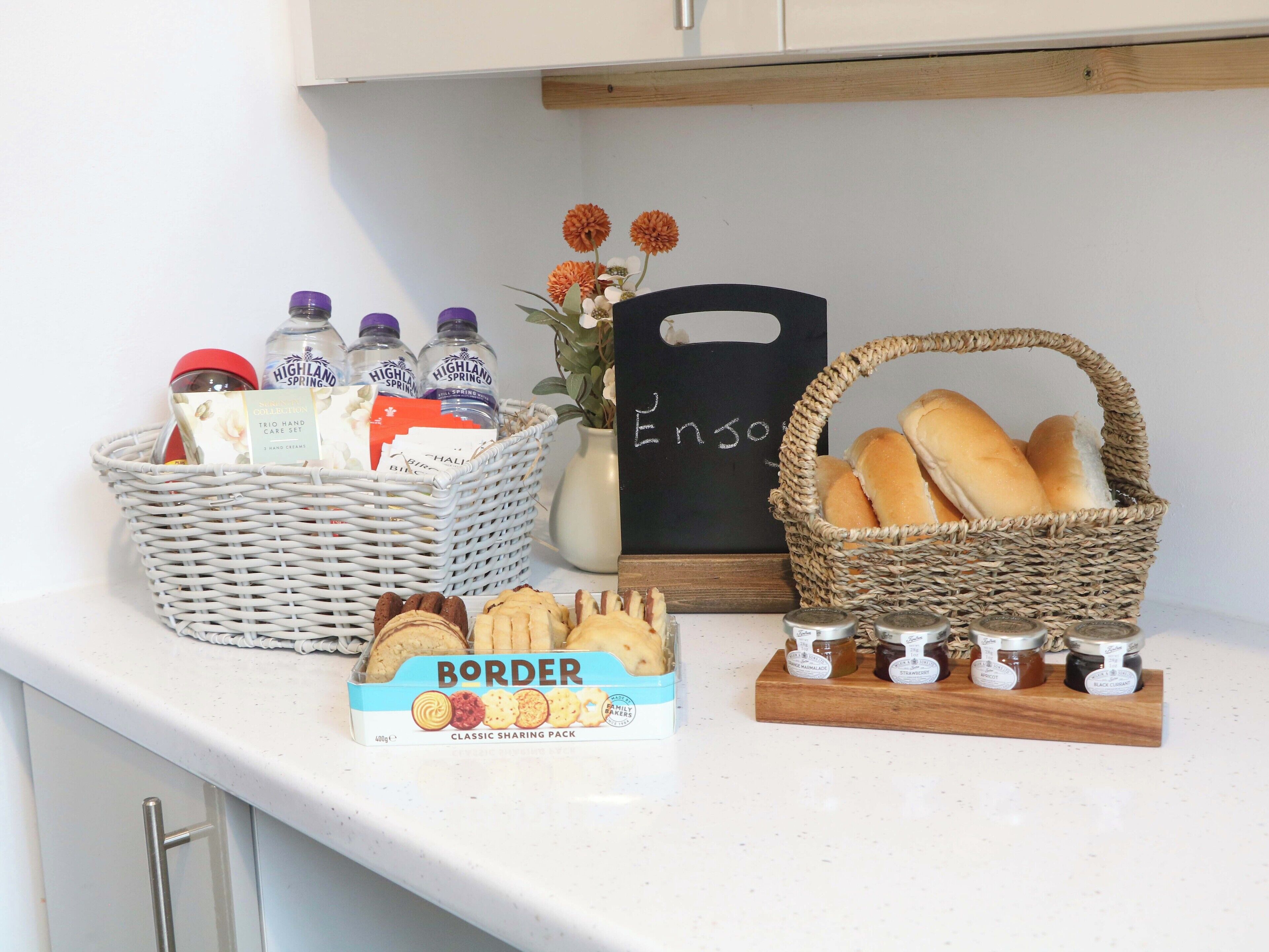 A kitchen counter with a basket of bread, a box of biscuits, a jar of jam, and a bottle of milk.