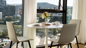 Uma sala de jantar com uma mesa posta para dois, um vaso com flores e vista para o horizonte urbano através de grandes janelas.
