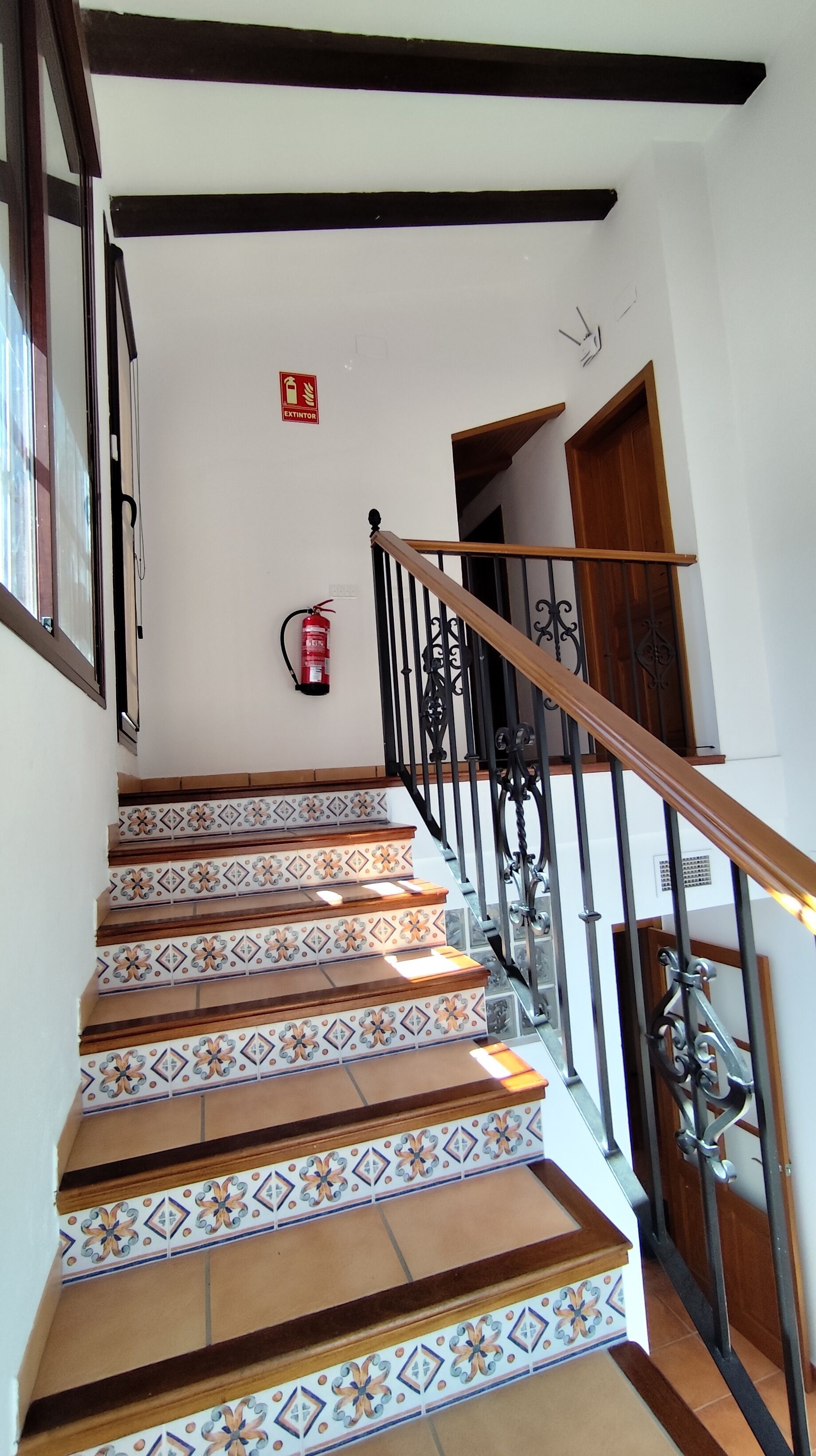 Una escalera con azulejos estampados, barandilla de madera y extintor en la pared.