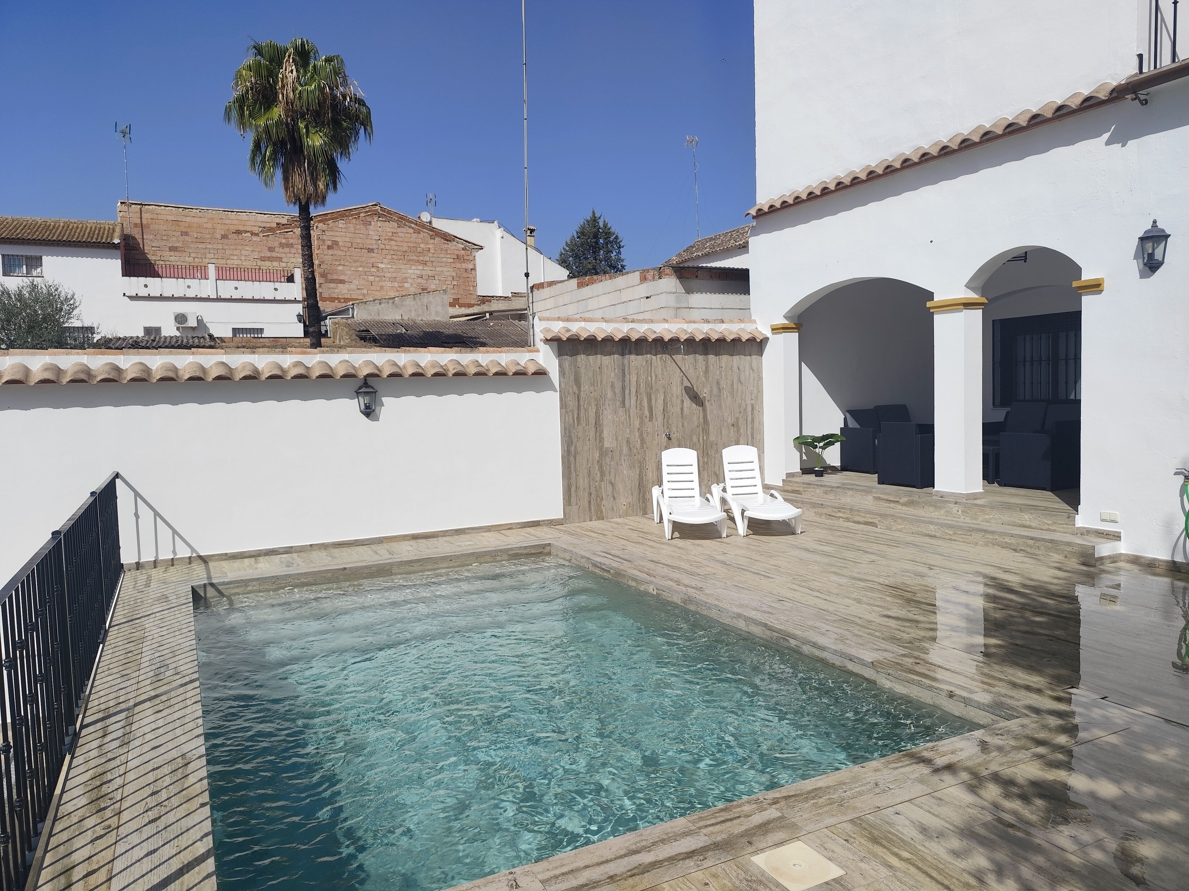 A swimming pool with two white lounge chairs, a wooden deck, and a white building with a tiled roof and arches.