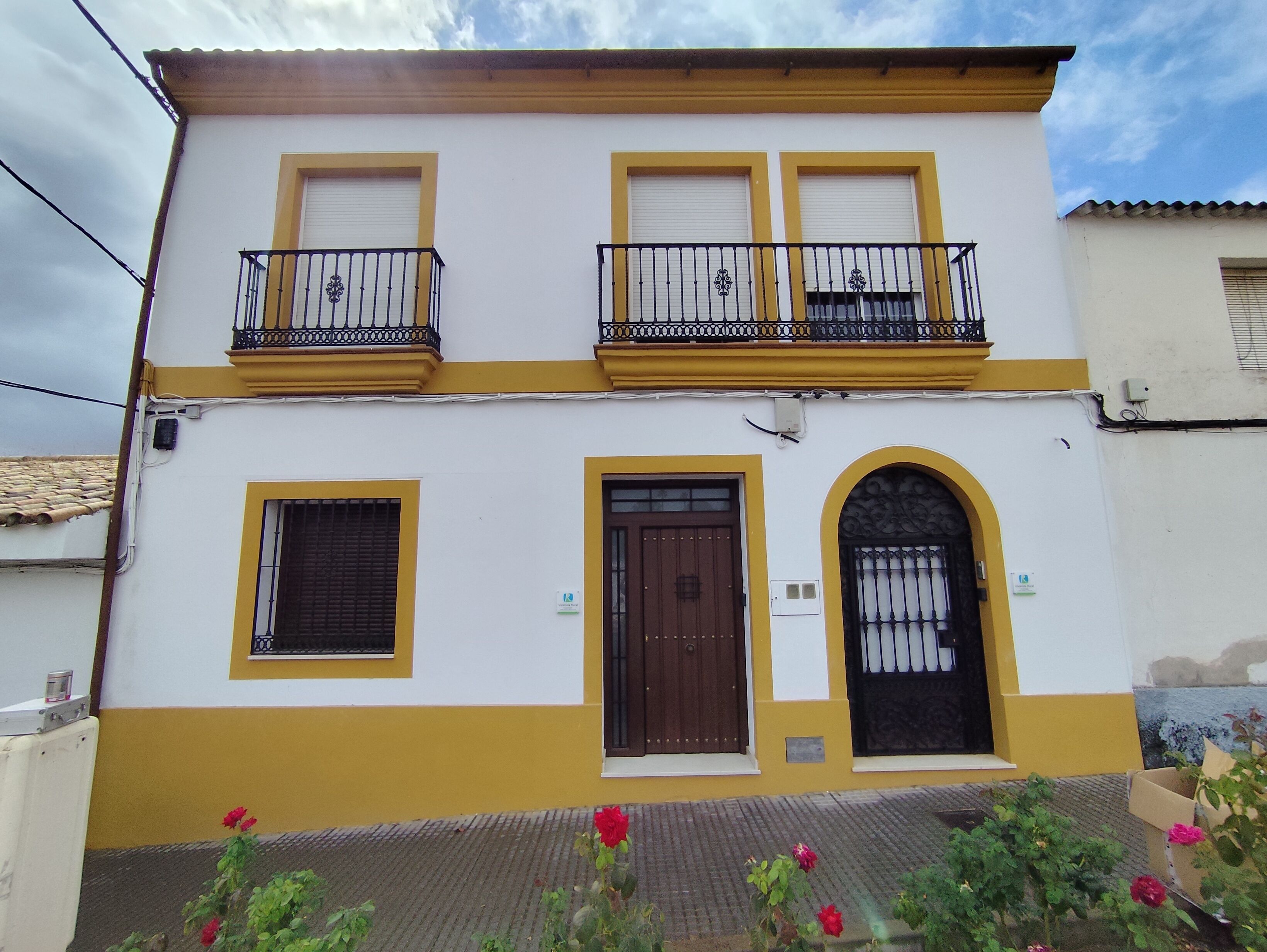 A two-story building with a yellow and white facade, featuring a central entrance with a brown door, two windows with shutters, and a balcony with black railings.