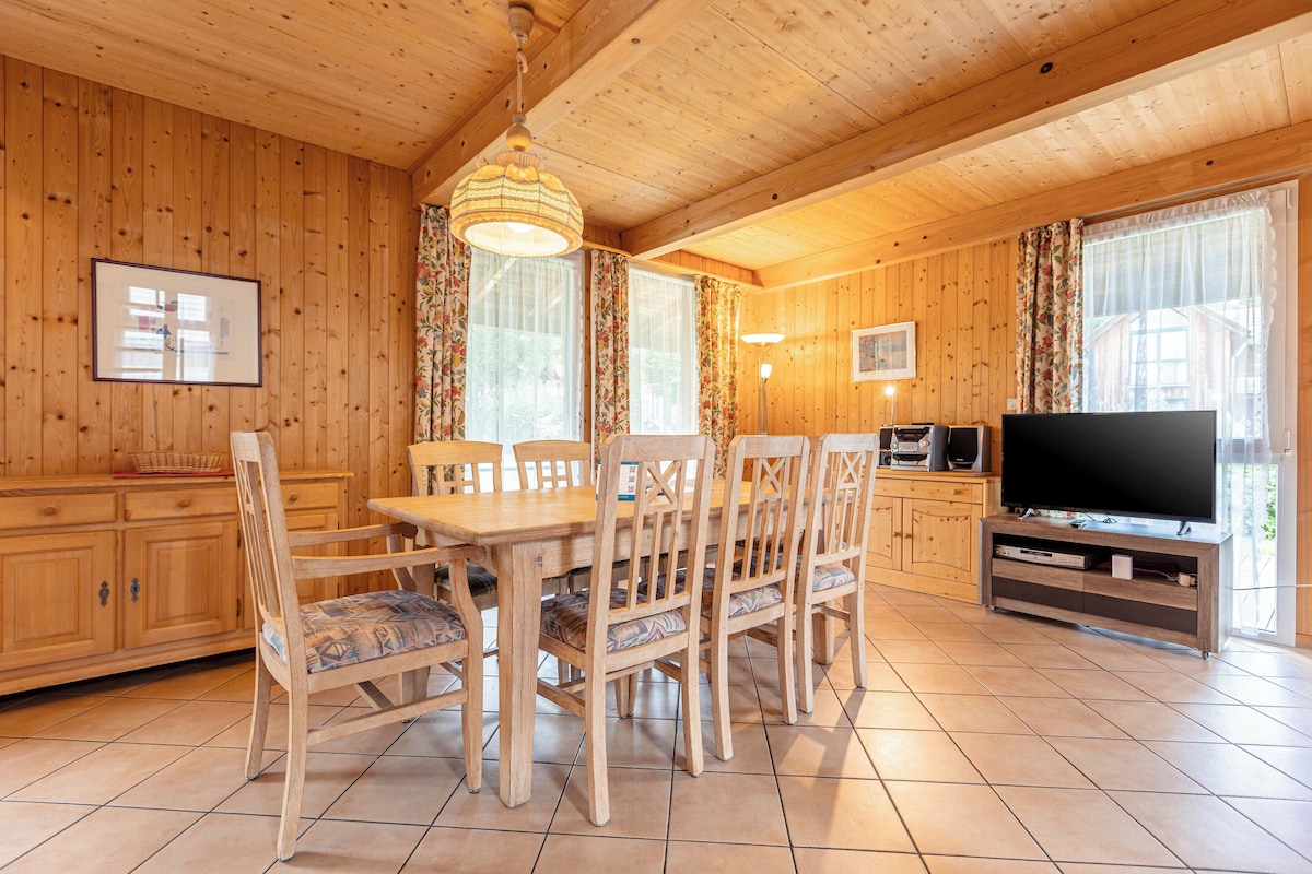 A wooden-paneled room with a dining table, chairs, a television, and a kitchen area.