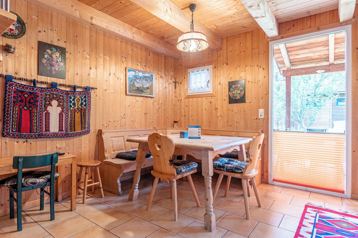 A wooden interior with a dining table, chairs, and a bench. There are framed pictures on the walls and a chandelier hanging from the ceiling.
