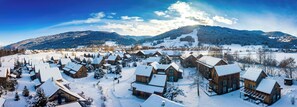 Vila coberta de neve com cabanas de madeira e montanhas ao fundo.
