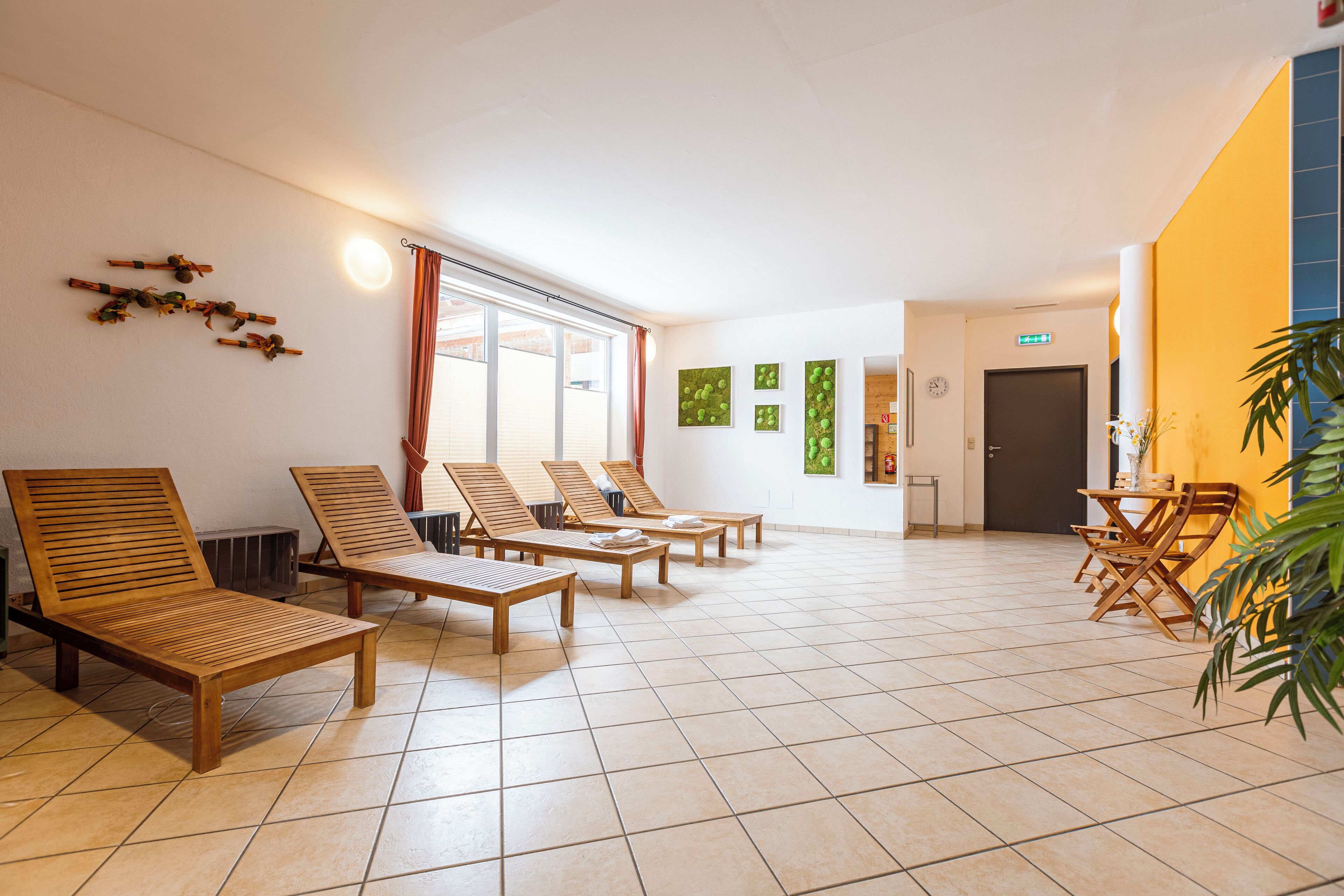 Ein geräumiges Zimmer mit Holz-Lounge-Stühlen und Fliesenboden, ein großes Fenster mit Vorhängen und eine Tür mit einem Notausgangsschild.