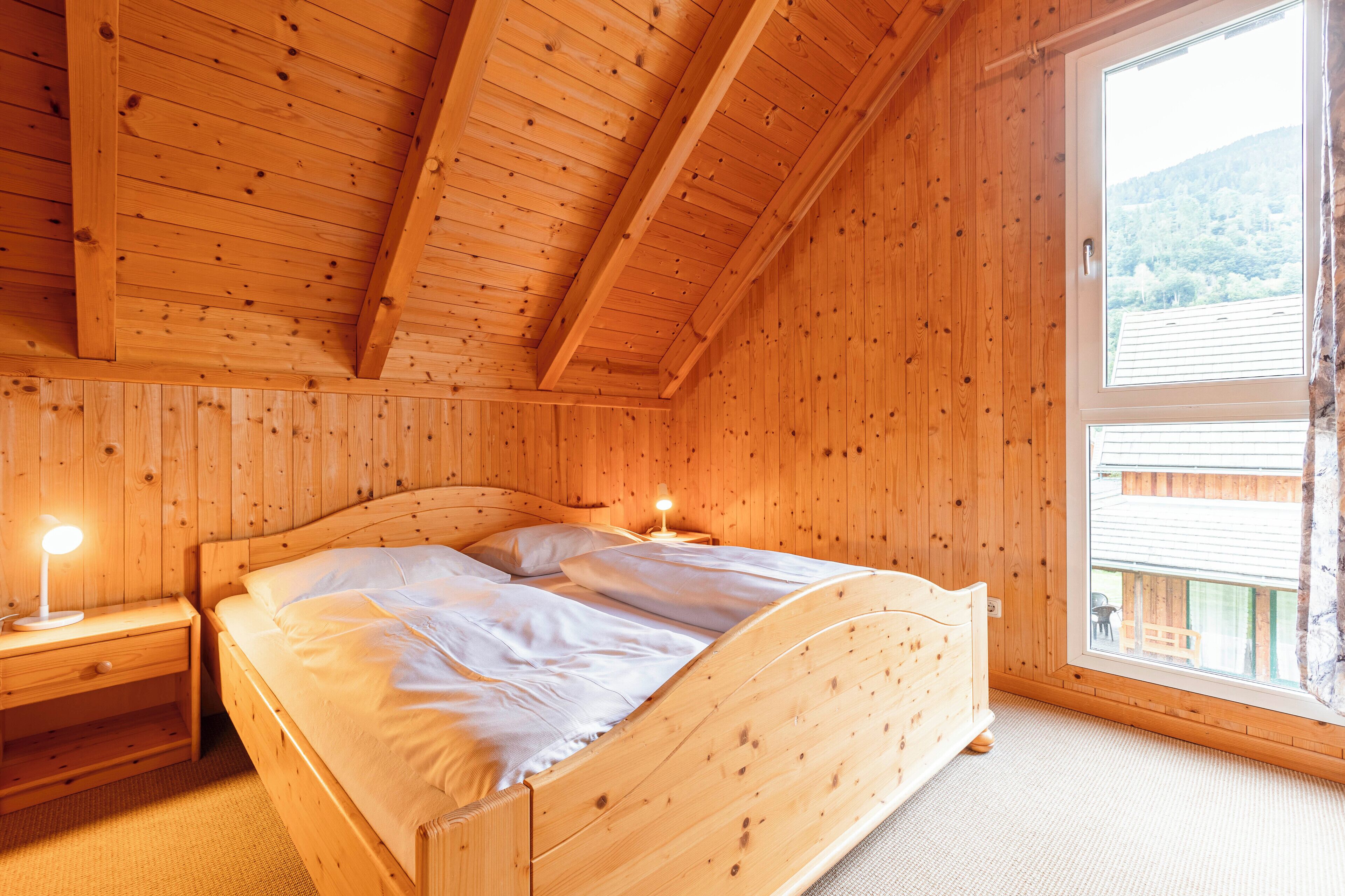 A wooden cabin bedroom with a bed, bedside tables, and a window with a view of a cabin and trees.