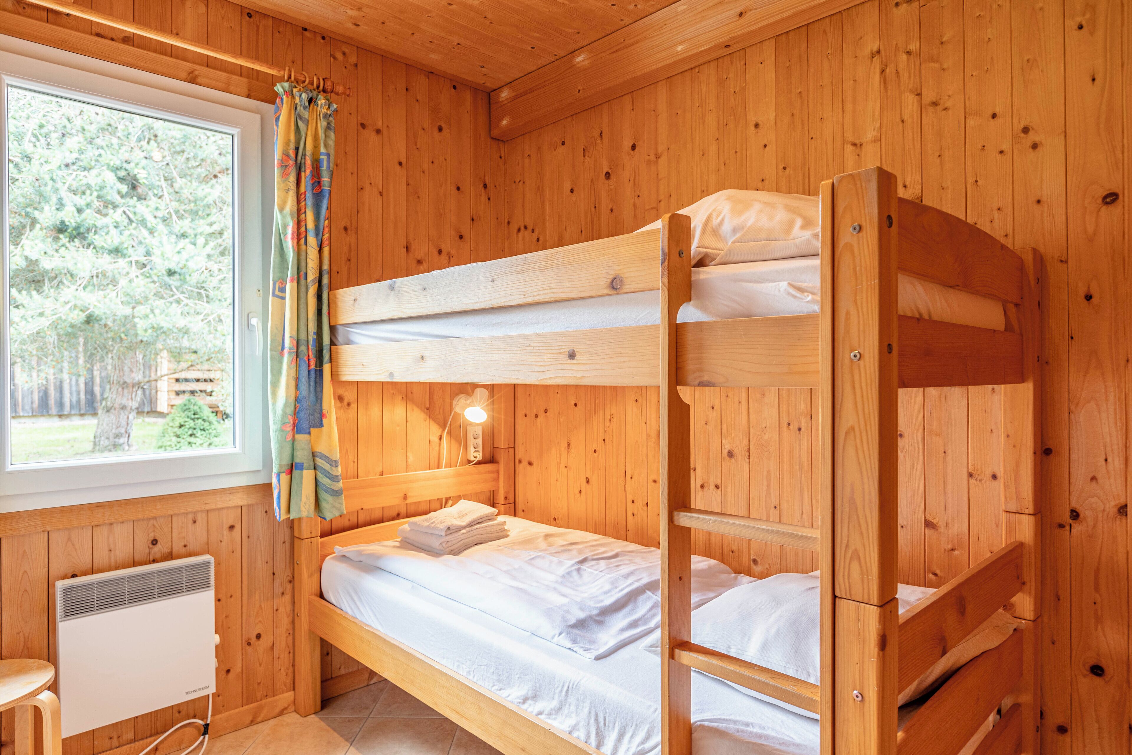 Habitación tipo cabaña de madera con literas, una ventana con cortinas, un calentador montado en la pared y una pequeña mesa.