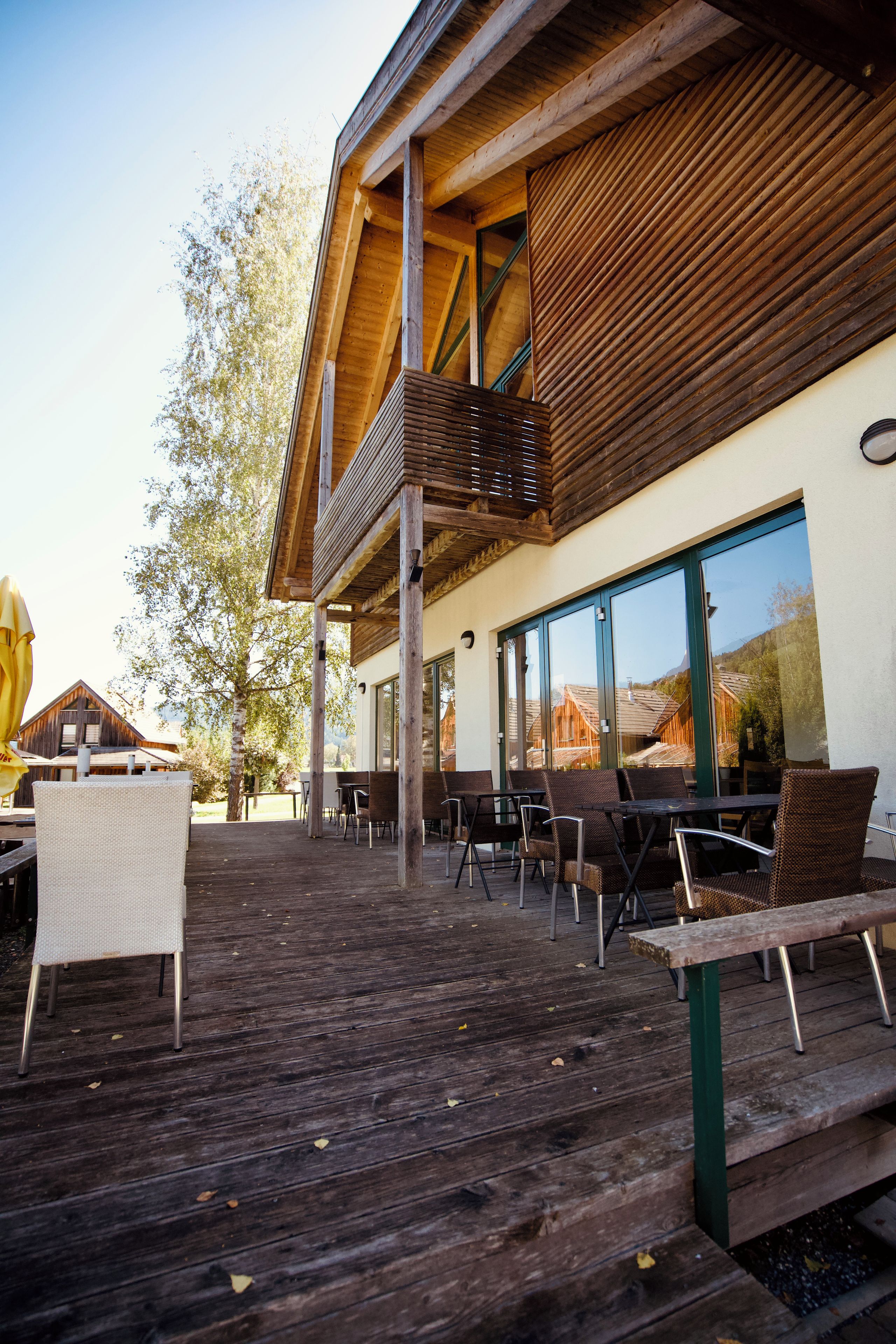 An outdoor seating area with wooden tables and chairs, a wooden deck, and a building with large windows.