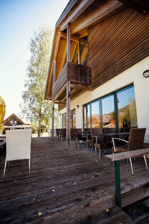 An outdoor seating area with wooden tables and chairs, a wooden deck, and a building with large windows. - FerienPark Kreischberg Ferienhäuser by ALPS RESORT (Sankt Lorenzen ob Murau)