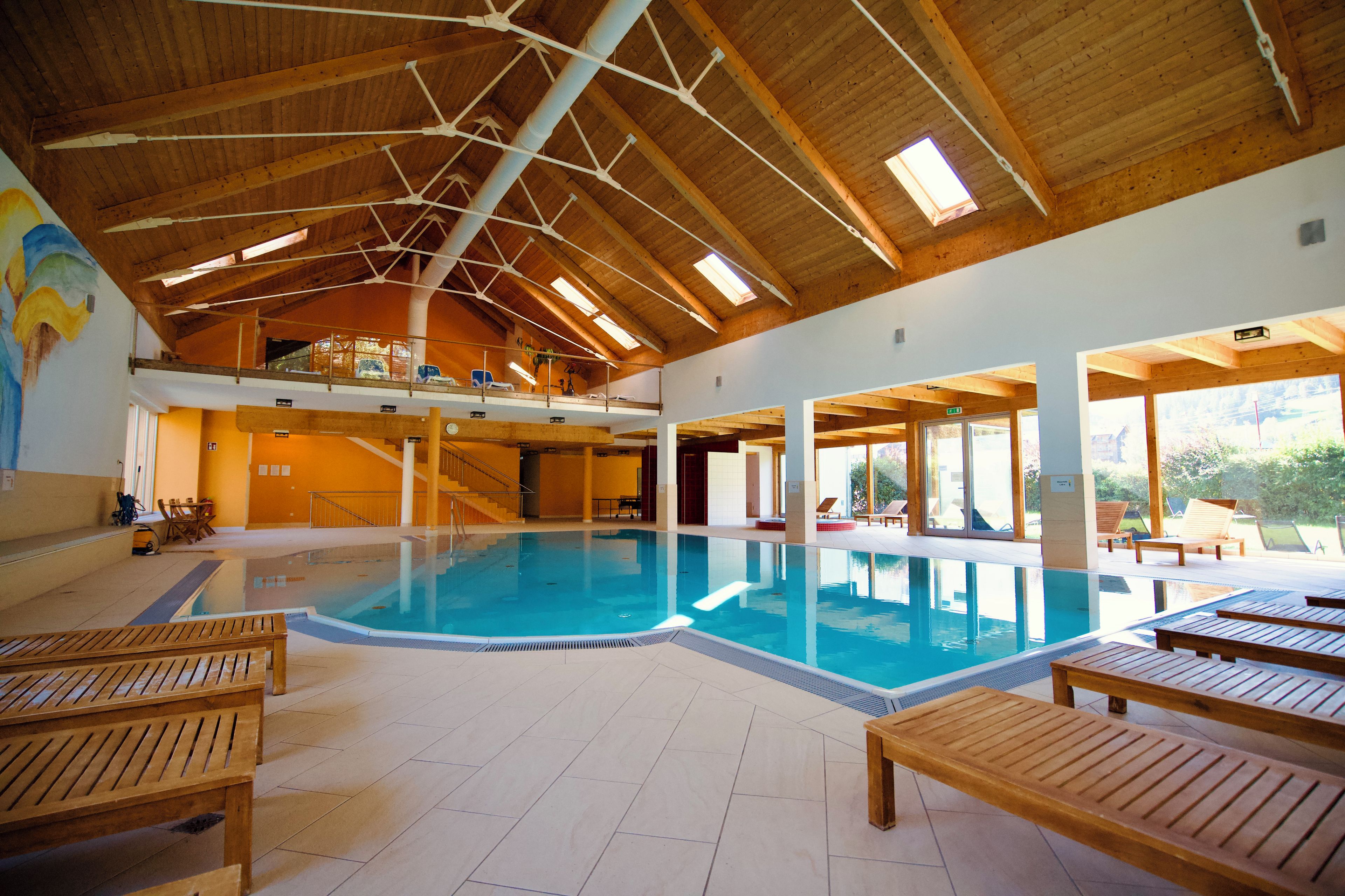 Une piscine intérieure dotée de chaises longues en bois, d’un haut plafond en bois avec des puits de lumière et offrant une vue sur l’extérieur.
