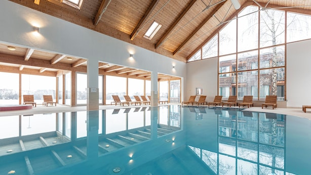 An indoor swimming pool with wooden beams and large windows, lounge chairs, and a view of the outside. - FerienPark Kreischberg Ferienhäuser by ALPS RESORT (Sankt Lorenzen ob Murau)