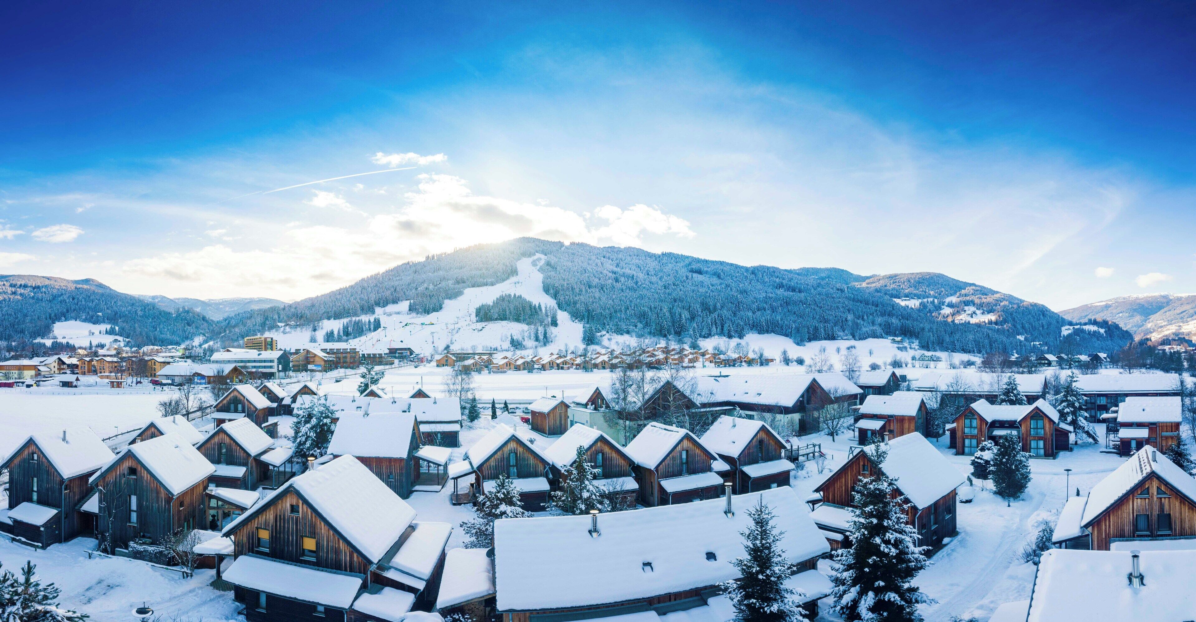 Un village enneigé avec des chalets en bois, un télésiège et des montagnes en arrière-plan.