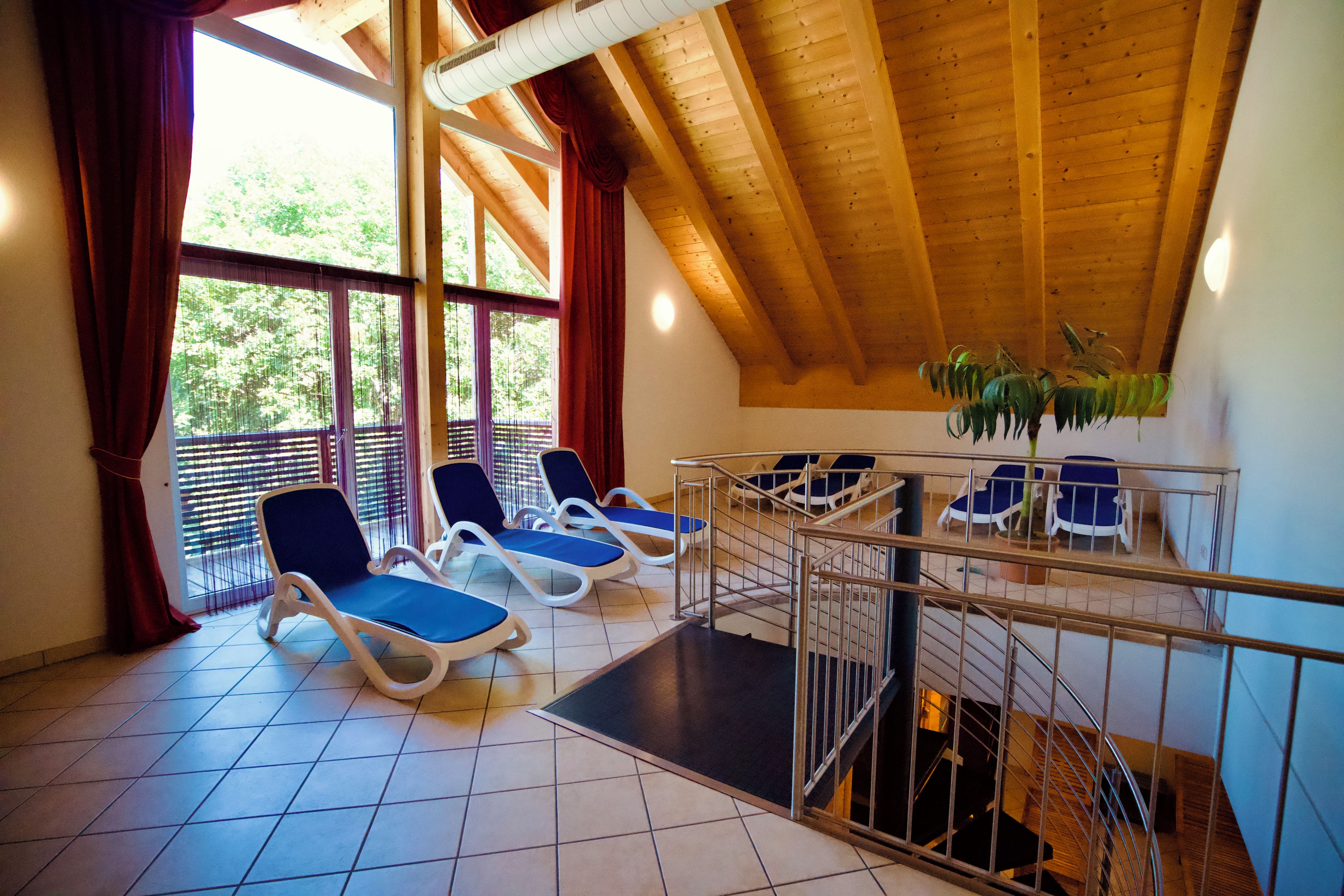 Een zonnige kamer met een houten plafond en uitzicht op groen. Er staan vier lounge fauteuils met blauwe kussens.