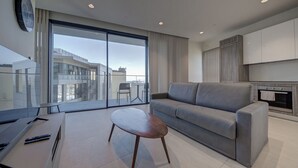A modern living room with a grey sofa, a small wooden table, and a large sliding glass door leading to a balcony with a city view. - Mercury Towers Luxury Stay w/ Rooftop Pool & Views (St. Julian`s)