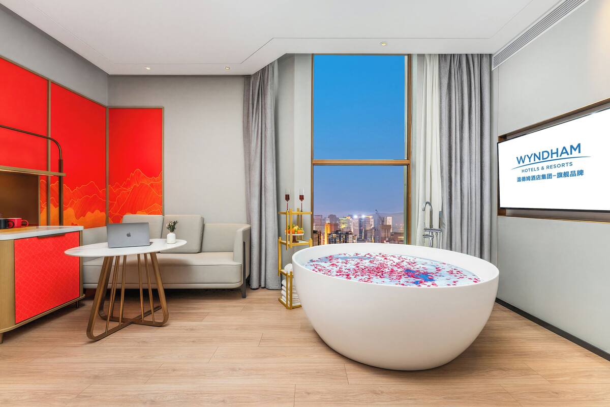 Ein modernes Hotelzimmer mit einer großen Badewanne, einem Laptop auf einem kleinen Tisch, einer Couch und Blick auf die Stadt.