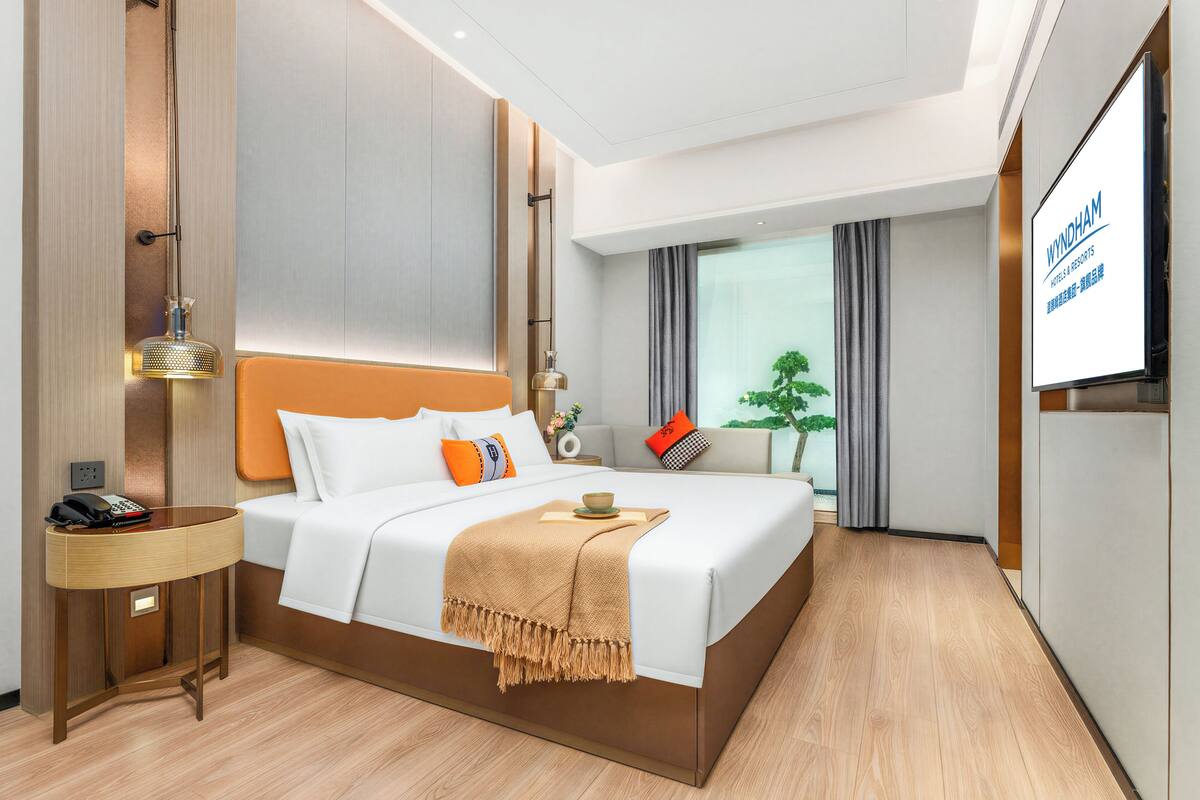 Ein modernes Hotelzimmer mit einem großen Bett, einem Nachttisch mit Lampe, einem Fernseher und Blick ins Freie.