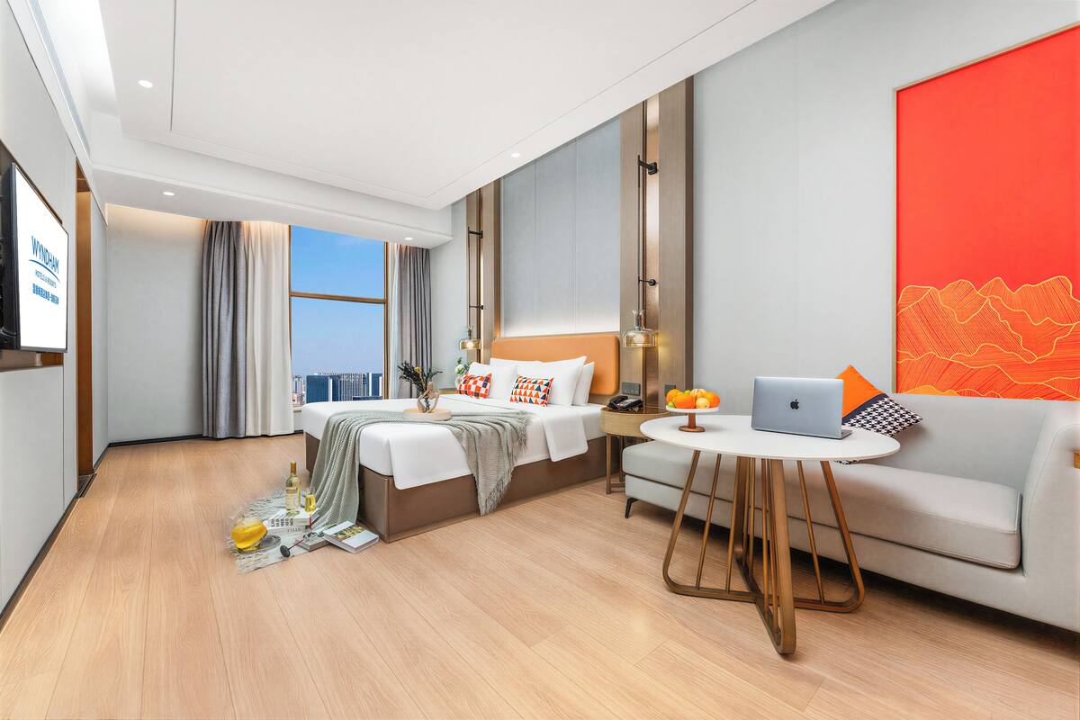 Ein modernes Hotelzimmer mit Bett, einer Couch, einem kleinen Tisch mit einem Laptop und einem großen Fenster mit Vorhängen.