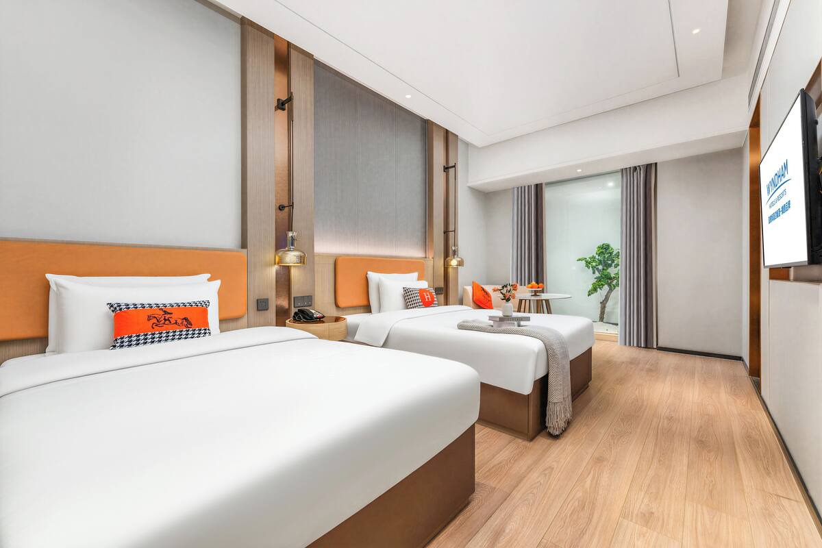 Ein Hotelzimmer mit zwei Betten, einem kleinen Tisch, einem Sessel, einem Fernseher und einem Fenster mit Vorhängen.