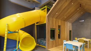 Un'area giochi per bambini con uno scivolo giallo, strutture in legno e una zona pranzo con sedie e tavolo azzurri.
