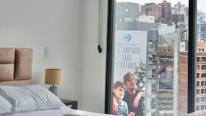 Una camera d'albergo con un letto, una finestra vista città, un quadro appeso al muro e un comodino con una lampada.