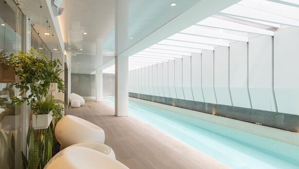 Un'area piscina interna moderna con soffitto in vetro, pareti bianche e sedute a bordo vasca.