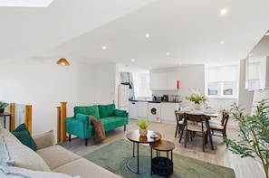 Un apartamento moderno con sala de estar, comedor y cocina. Incluye un sofá verde, una mesa de comedor con sillas y una cocina con gabinetes blancos y lavavajillas.