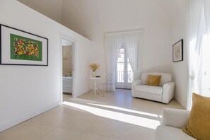 Amplio salón con un sofá blanco y una mesita con un florero, una silla blanca y un cuadro abstracto enmarcado en la pared.