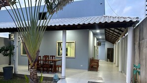 A covered patio area with a dining table and chairs, a small garden with a palm tree, and a concrete pathway leading to a building entrance. - BEACH HOUSE (Prado)