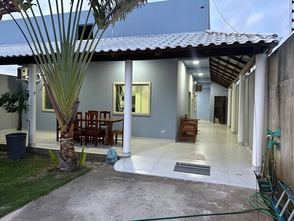 A covered patio area with a dining table and chairs, a small garden with a palm tree, and a concrete pathway leading to a building entrance. - BEACH HOUSE (Prado)