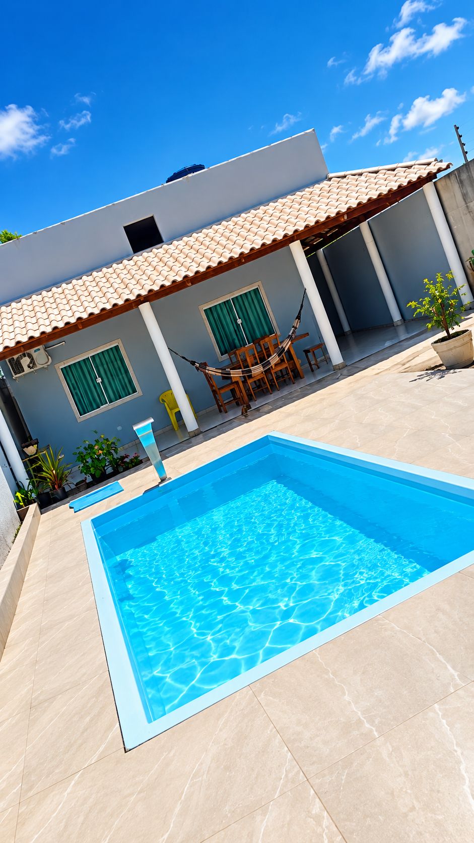 A swimming pool with a tiled deck, a covered patio area with chairs, and a building with a tiled roof.