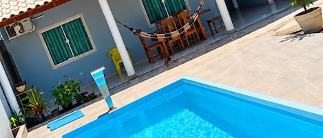 A swimming pool with a tiled deck, a covered patio area with chairs, and a building with a tiled roof.