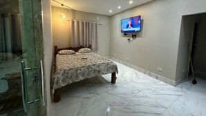 A bedroom with a bed, a TV mounted on the wall, and a bathroom with a glass shower enclosure. - BEACH HOUSE (Prado)