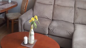 Olohuoneessa on beigenvärinen sohva, pyöreä sohvapöytä, jonka päällä on kynttilä ja maljakko kukkineen, puinen ruokapöytä, jonka päällä on myös maljakko kukkineen, sekä seinällä oleva maalaus, jossa on kuvattuna rantamaisema.