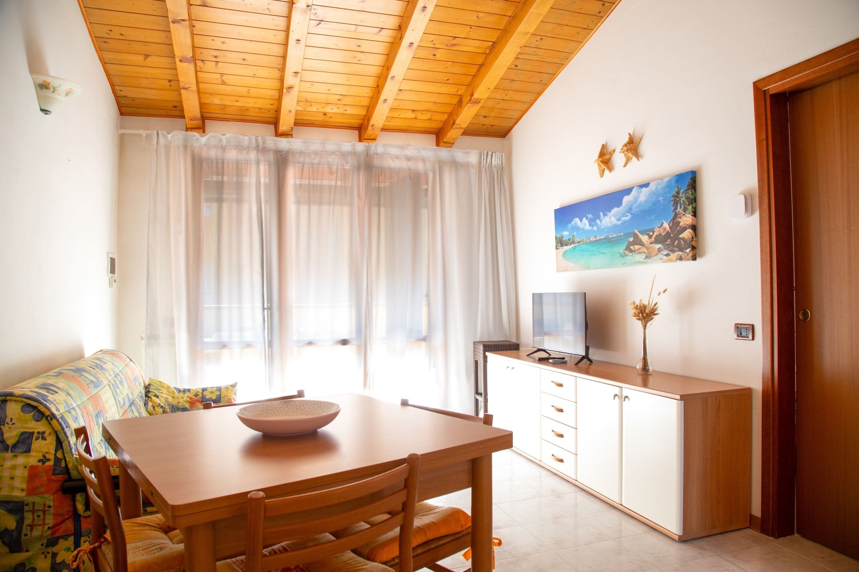 Um quarto aconchegante com uma mesa de jantar de madeira, um sofá com um cobertor colorido, um quadro de uma praia e um teto de madeira.