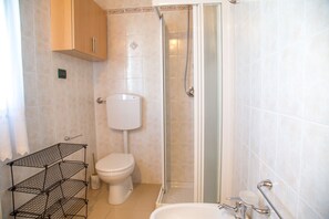 Egy fürdőszoba, amelyben WC, zuhanyzó, mosdókagyló és egy polcrendszer található.