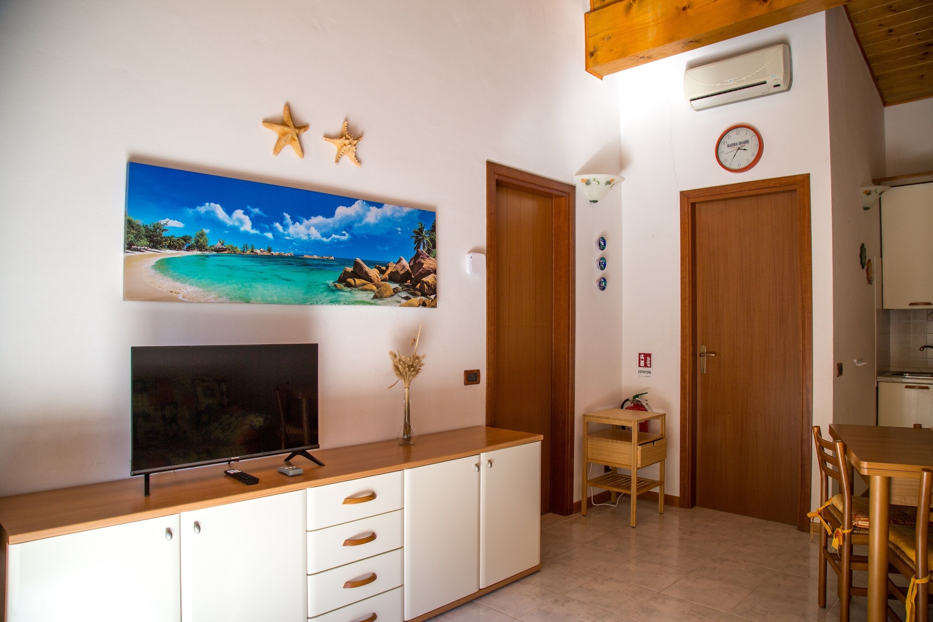 Um espaço de estar compacto com televisão em um armário de madeira, área de jantar com mesa e cadeiras, e cozinha com geladeira e fogão.