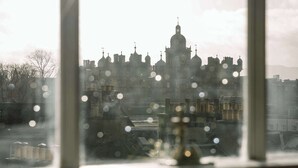 Visão através de uma janela, refletindo um horizonte urbano, com um edifício imponente com cúpula e várias torres.