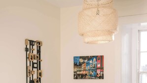 Uma sala de estar moderna com um sofá cinza, uma luminária pendente trançada, uma obra de arte na parede e uma janela com vista para edifícios.