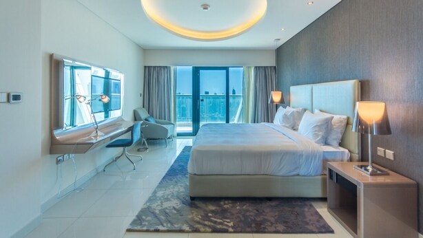A modern hotel room with a large bed, a desk, a chair, and a view of the city through large windows. - Spacious 1BR Rental | 1050 sq ft | Luxury Lifestyle in Paramount Towers (Dubai)
