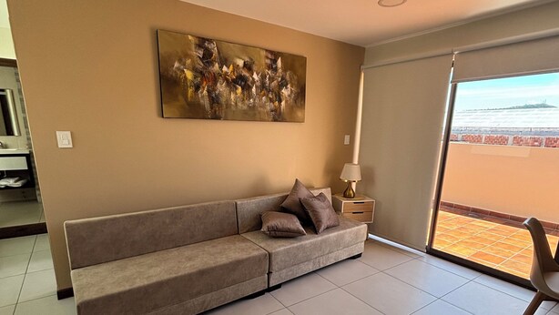 A modern living room with a beige sofa, a large abstract painting, a floor lamp, and a view of a distant landscape through a sliding door.