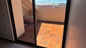 A room with a sliding glass door leading to a balcony with a tiled floor and a view of buildings and a clear sky.