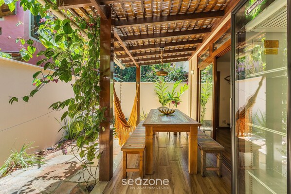 A covered patio with a wooden table and benches, surrounded by plants and a glass door.