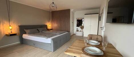 A modern bedroom with a large bed, bedside lamps, and a dining area with a wooden table set for two.