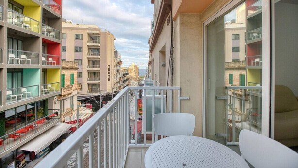 Un balcon avec des chaises blanches et une table, donnant sur une rue bordée de bâtiments aux couleurs vives et sur laquelle se trouve un restaurant en contrebas.