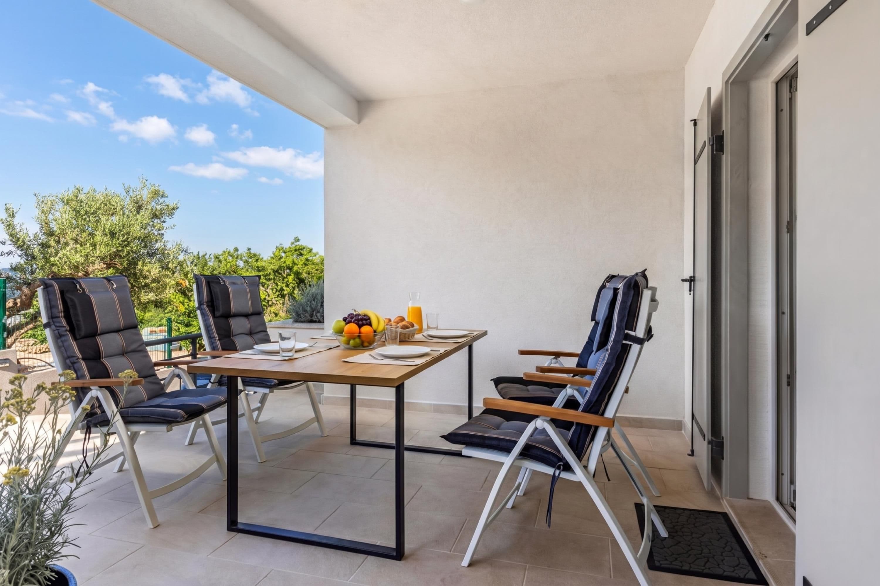 A balcony with a table set for two, two chairs, a fruit bowl, and a bottle of orange juice.