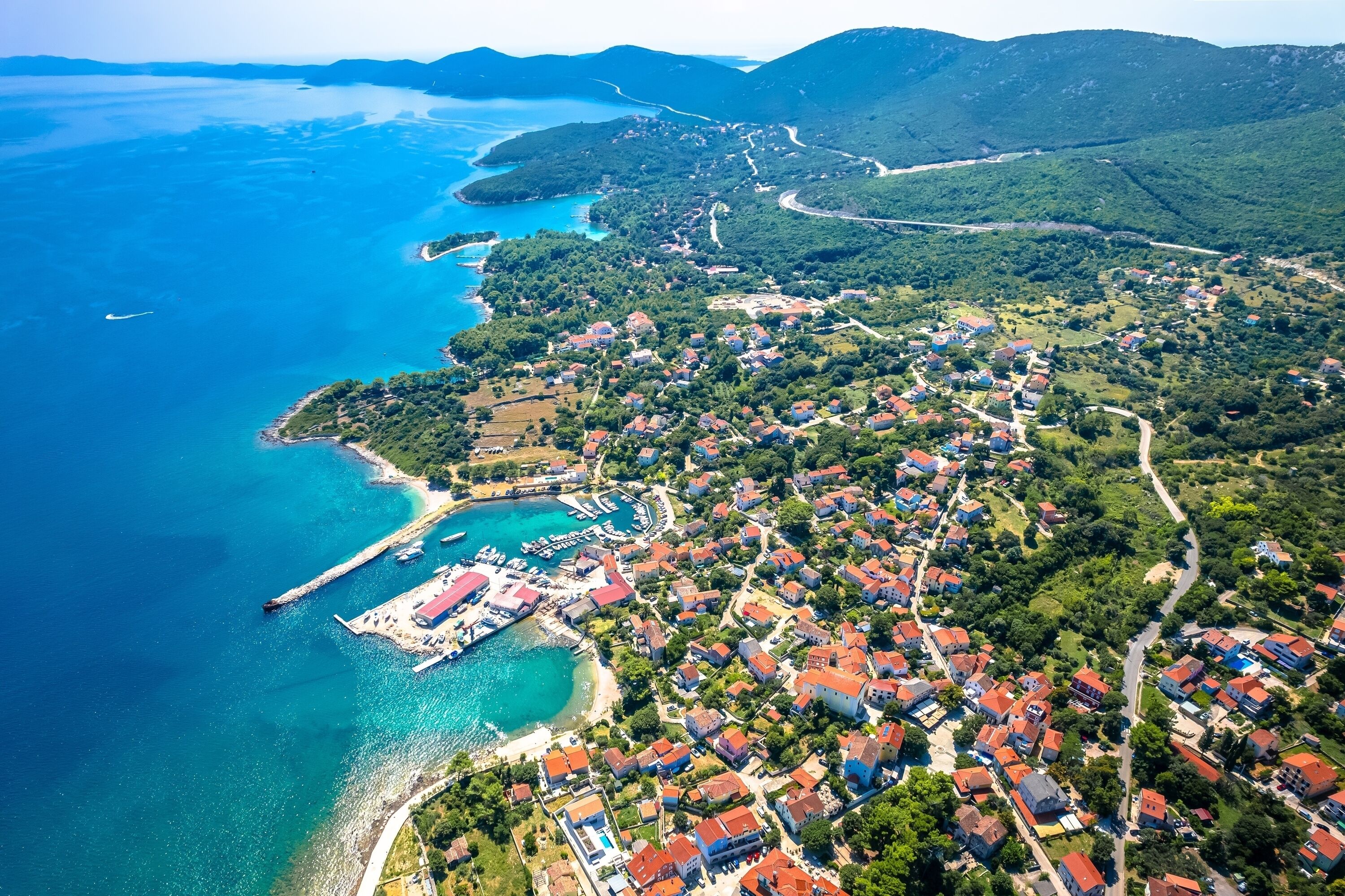 An aerial view of a coastal town with a marina, residential areas, and surrounding hills.