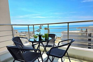Een balkon met een tafel en stoelen, uitzicht op zee, en gebouwen op de achtergrond.